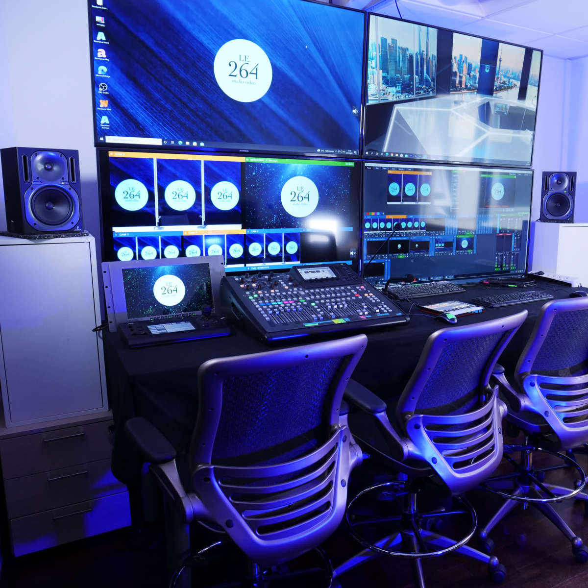 A modern control room with multiple large screens, audio equipment, and rows of swivel chairs arranged in front of a desk.