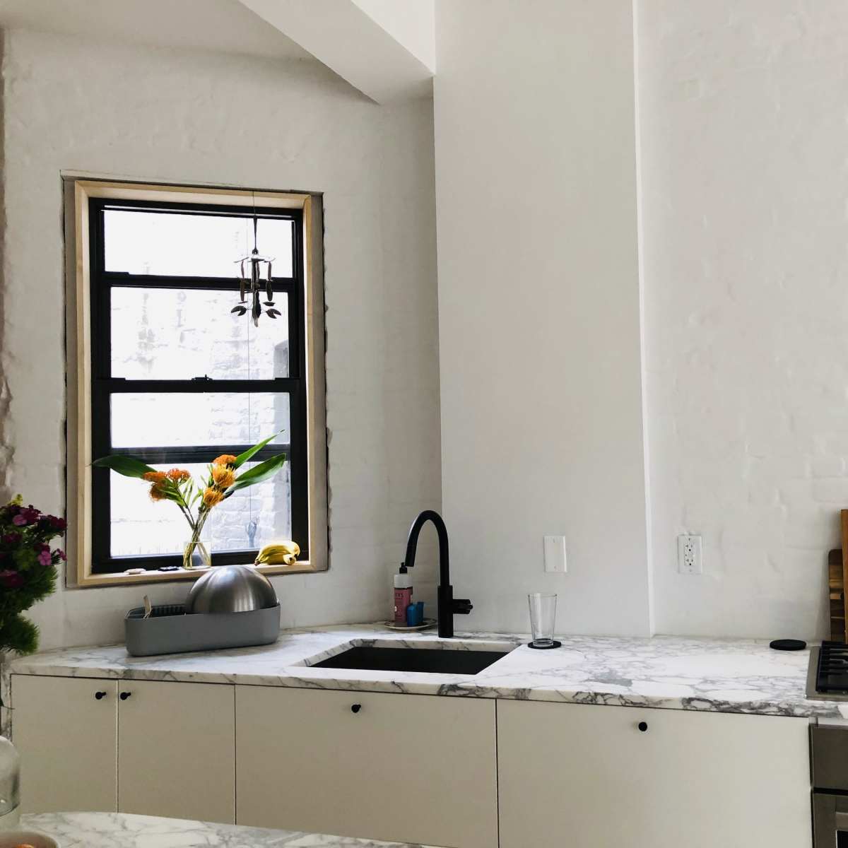 A modern kitchen features a marble countertop, a sink with a black faucet, a gas stove, and a window displaying a plant arrangement.