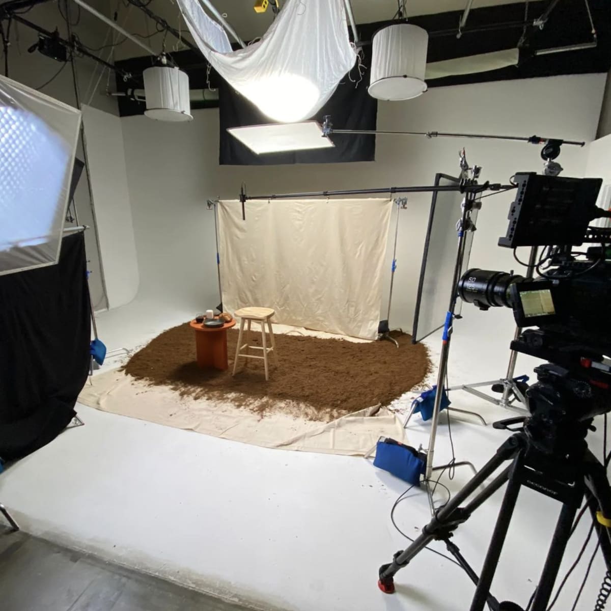 A photography studio is set up with lights and a camera, featuring a bare white backdrop and a stool placed on a carpet of brown material.