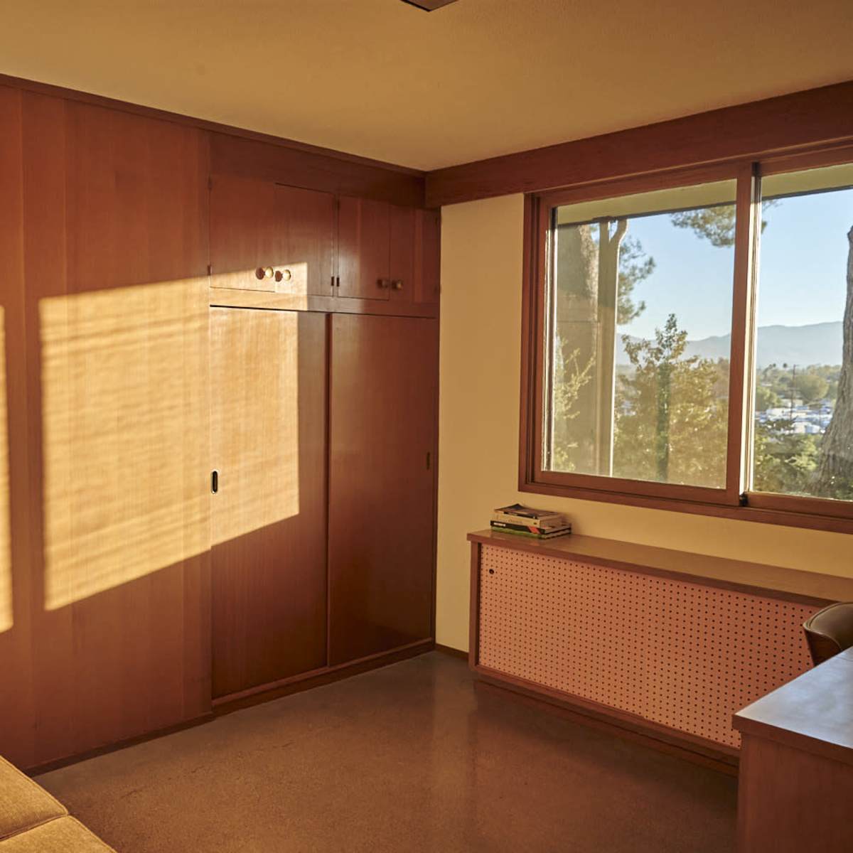 A room features wooden walls, a window with sunlight casting shadows, and minimal furnishings including a sofa and a cabinet.
