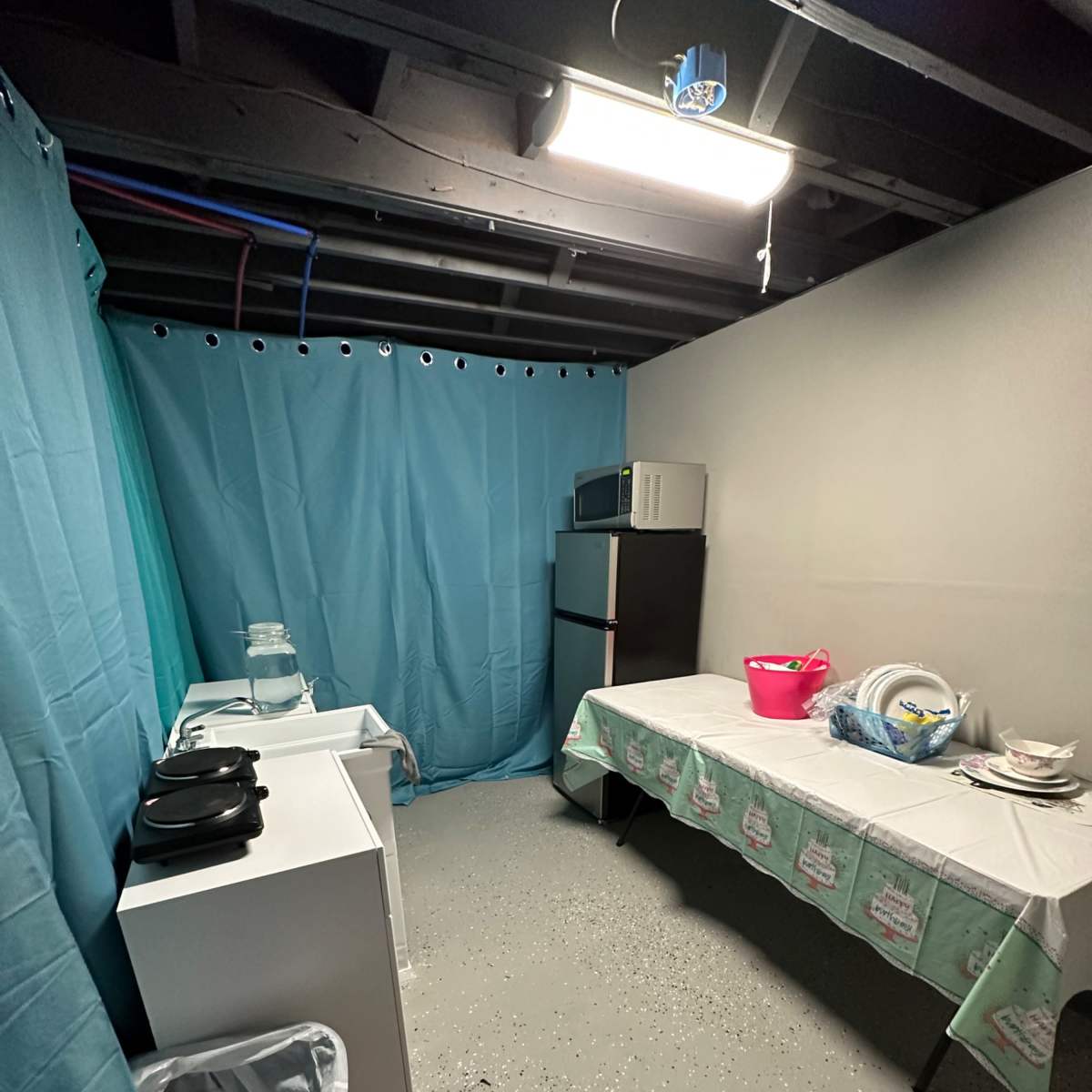 A small kitchen area in a basement, featuring a table with dishes, a microwave, a refrigerator, and a trash can, all surrounded by light blue curtains.
