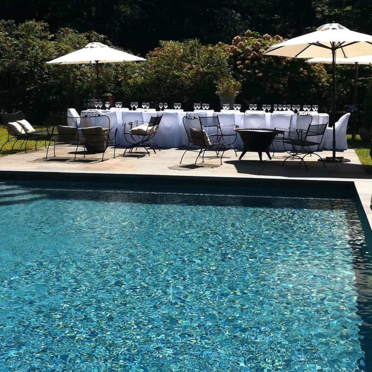 A long table set for dining with glasses sits beside a swimming pool and under umbrellas, surrounded by green foliage.