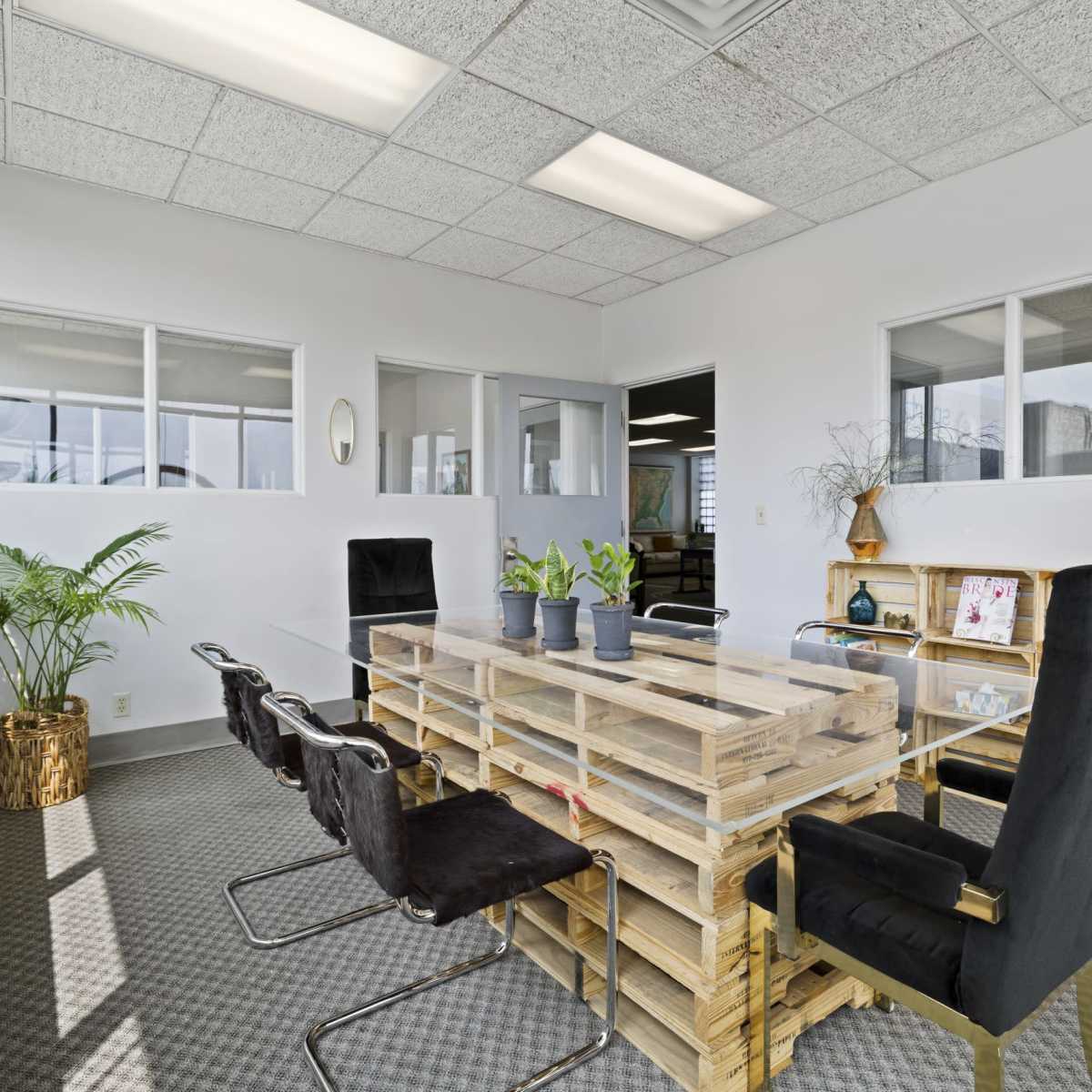 A modern conference room with a large pallet wood table surrounded by black chairs and plants near the windows.