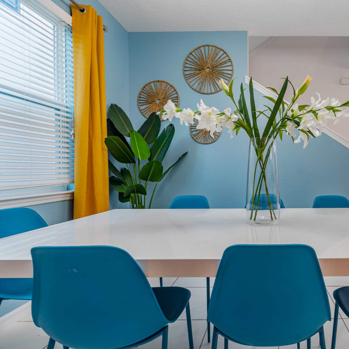 A modern dining area features a white table surrounded by blue chairs, with a vase of white flowers and a backdrop of blue walls and a yellow curtain.