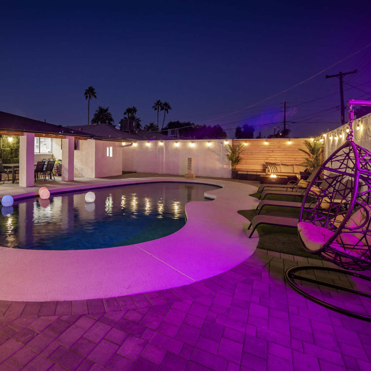 A desert backyard at night featuring a swimming pool with colored lights, lounge seating, and palm trees illuminated by string lights.