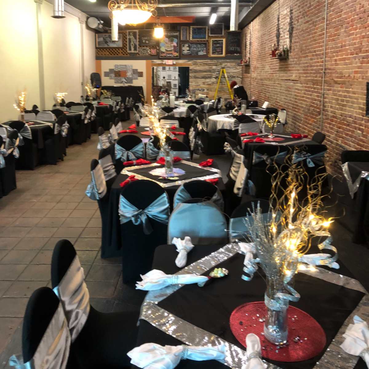 A spacious dining area set with numerous tables covered in black cloth, adorned with silver and blue decorations, and featuring centerpieces with lights.