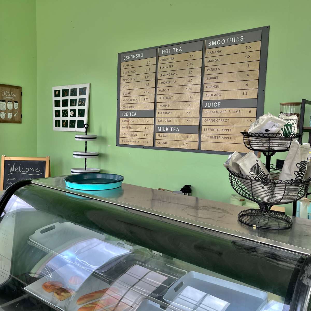A cafe interior with a green wall, a refrigerated display case, and a menu board listing various beverages and snacks.