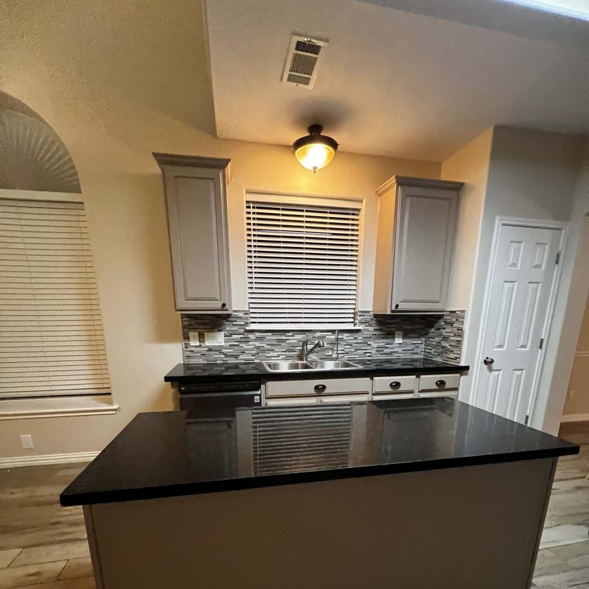 A kitchen features light gray cabinets, a black countertop, and a tiled backsplash with a window and blinds above the sink.