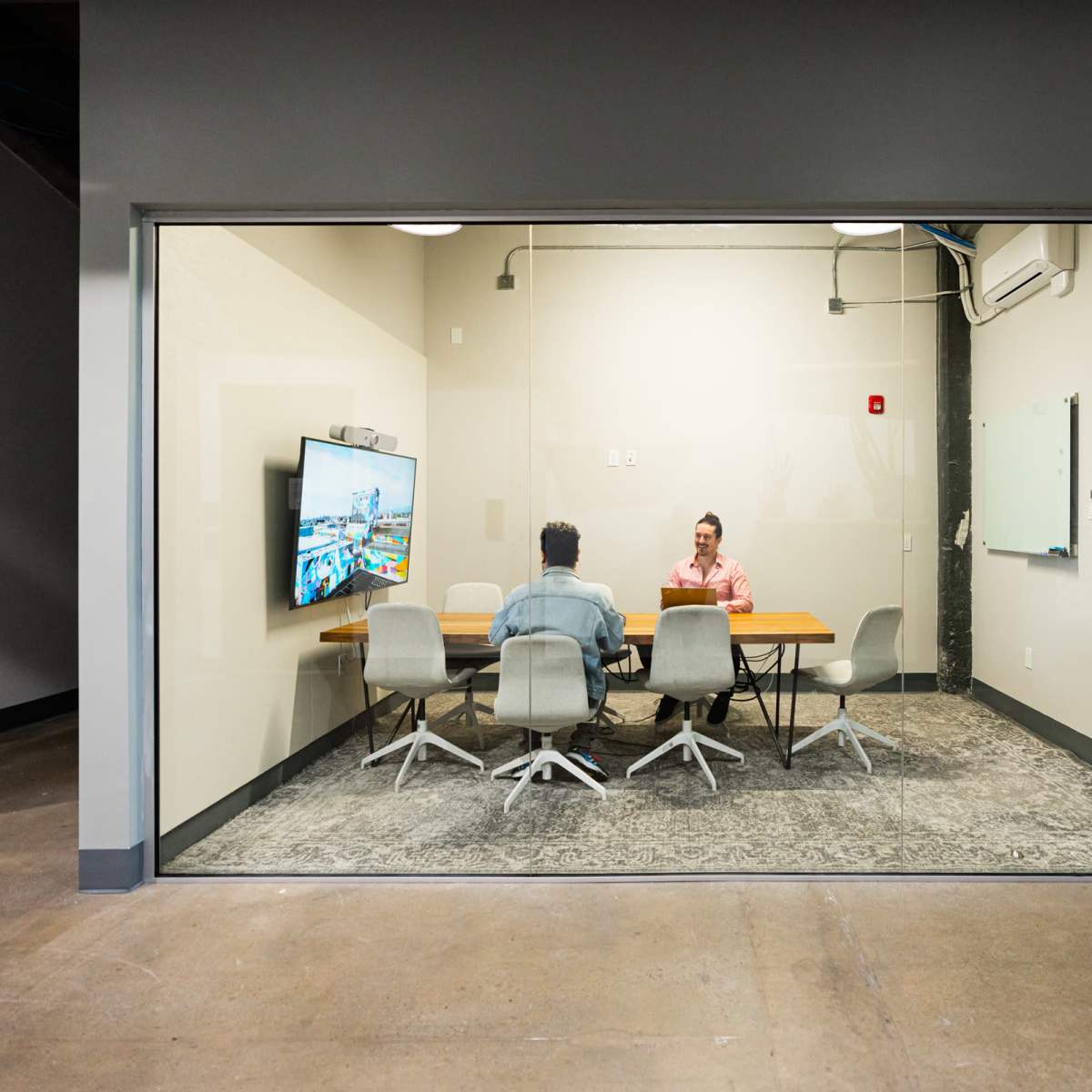 Two people are seated at a conference table in a glass-walled meeting room, with a large screen displaying a digital presentation.