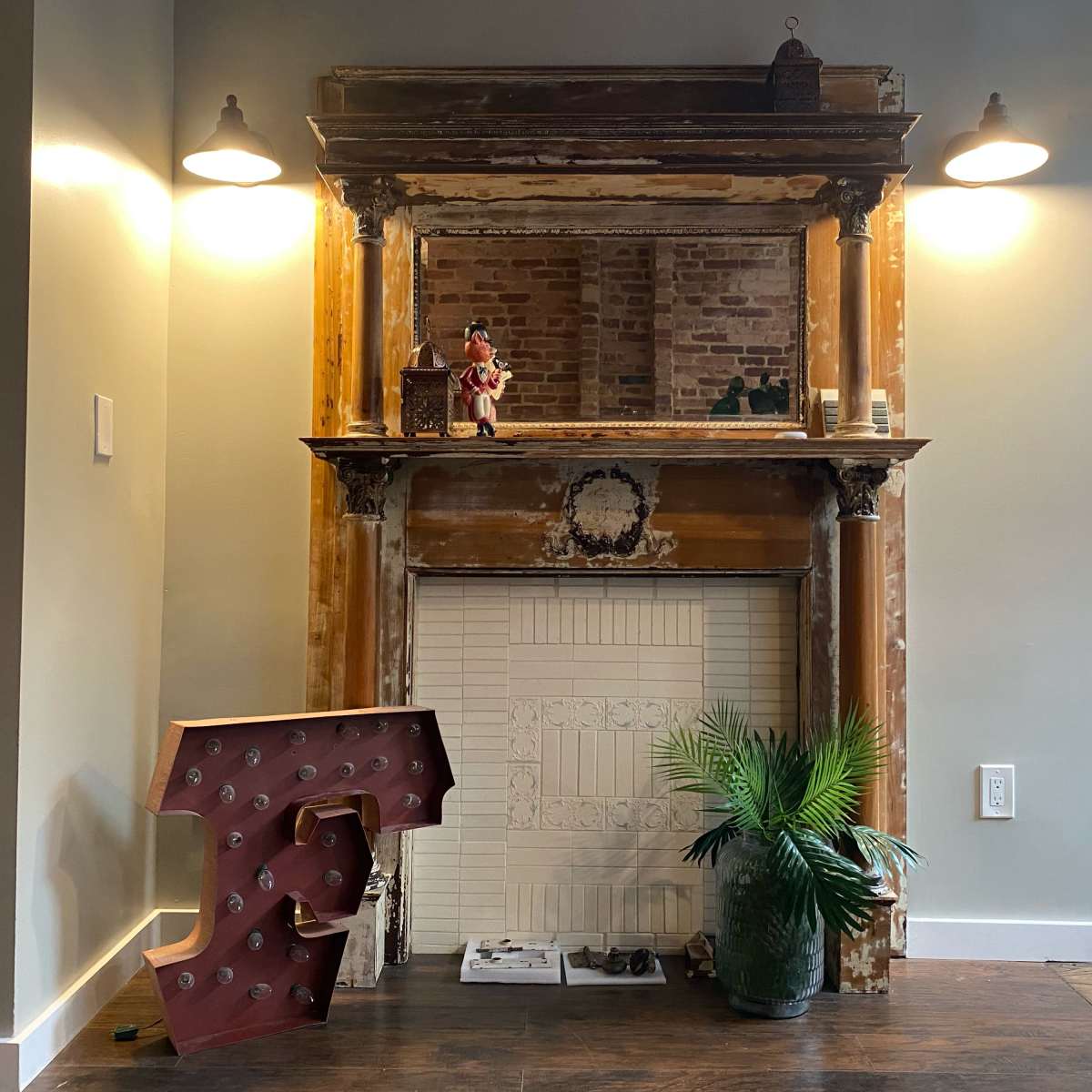An ornate fireplace mantel with a decorative mirror above it, flanked by two wall sconces, and a large, red, illuminated number five positioned beside a potted plant on the floor.
