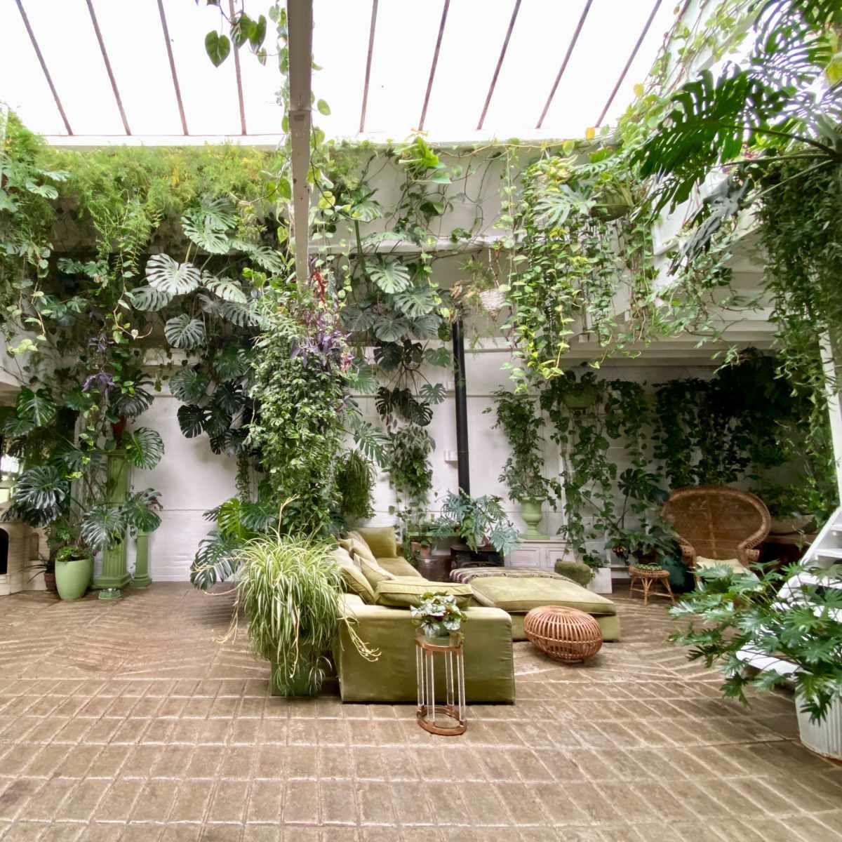 A spacious, indoor area filled with various green plants, featuring a large sofa, wicker furniture, and a staircase leading to an upper level.