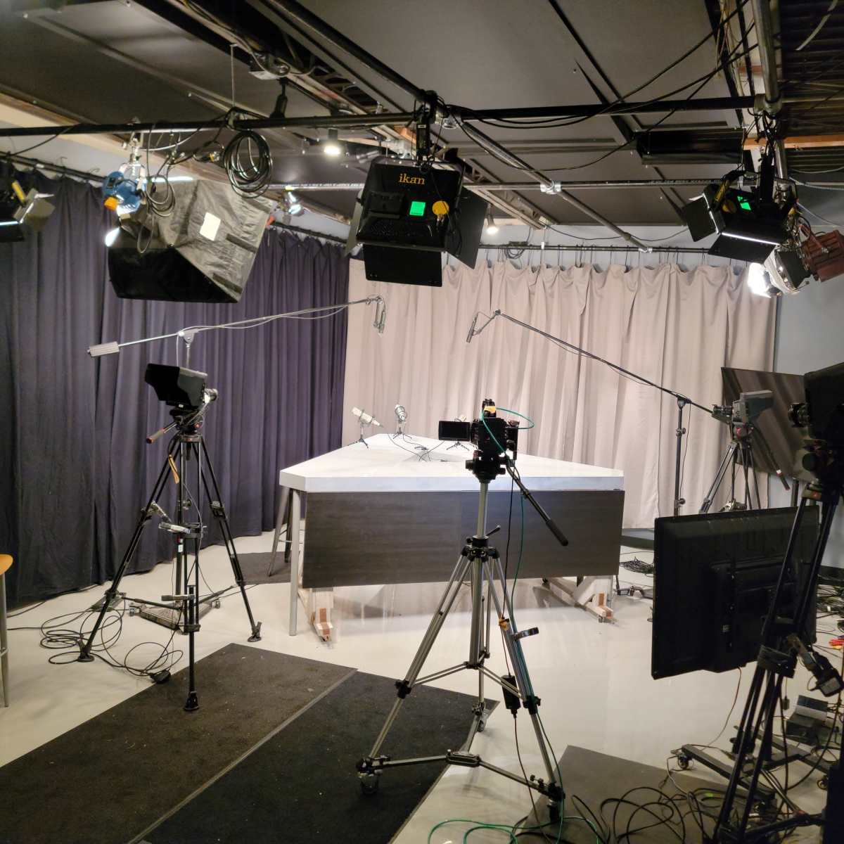 A television studio setup features several cameras, lights, and a central table with a backdrop of curtains.