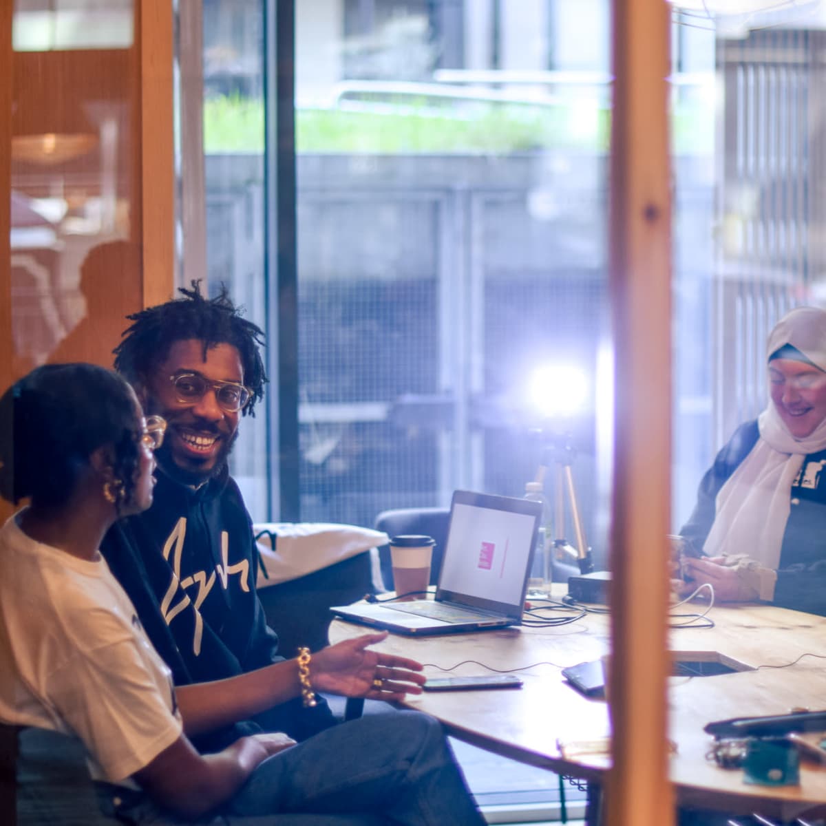 A group of three people is engaged in a conversation around a table while a fourth person sets up a camera in a brightly lit room.