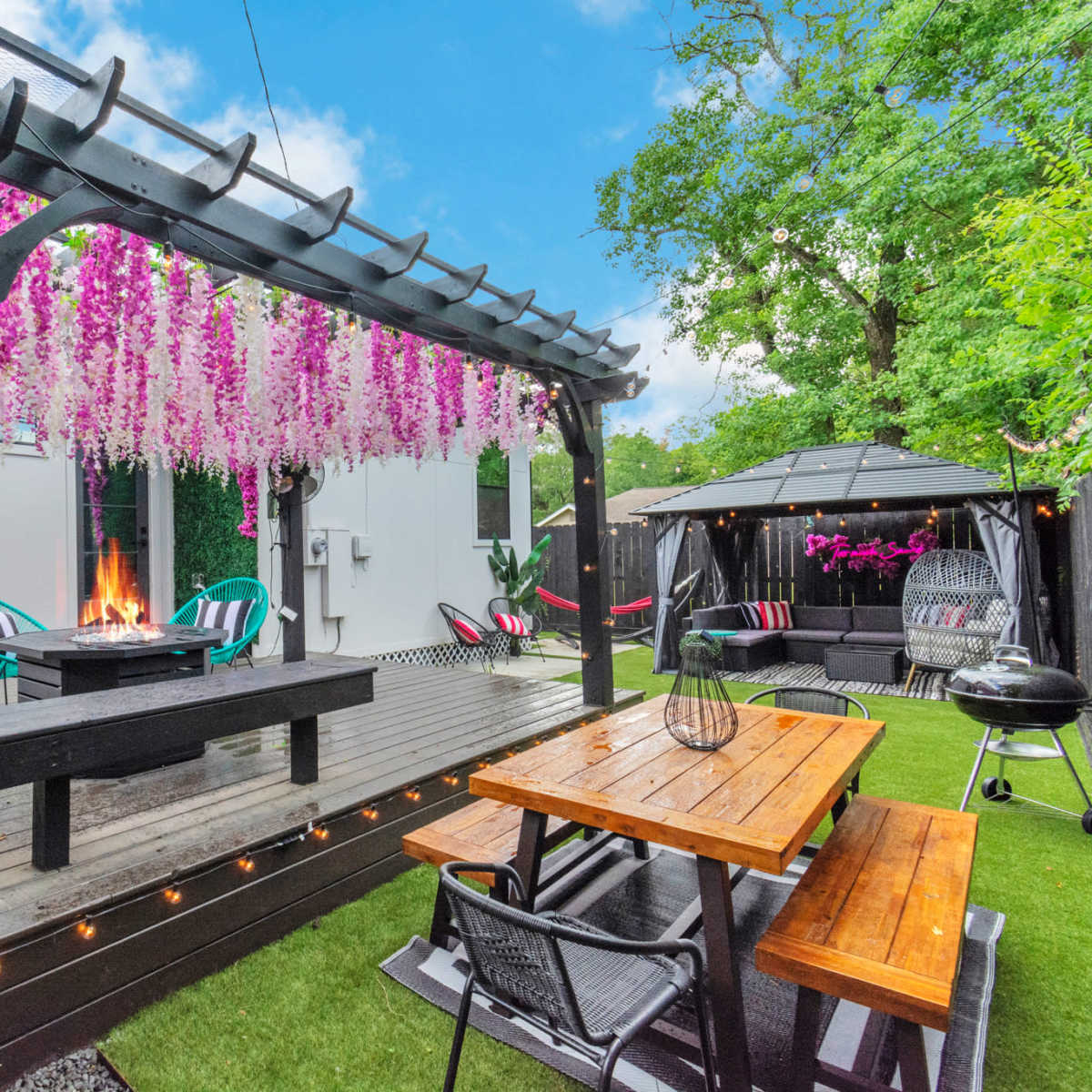 A backyard with a wooden deck featuring a fire pit, surrounded by seating, vibrant hanging flowers, and a gazebo in a well-landscaped garden.