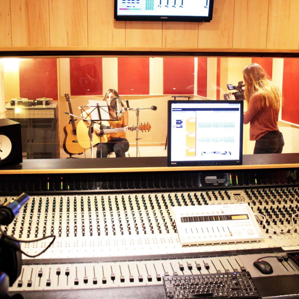 A musician plays an acoustic guitar in a recording studio while being filmed, with a sound mixing console in the foreground.