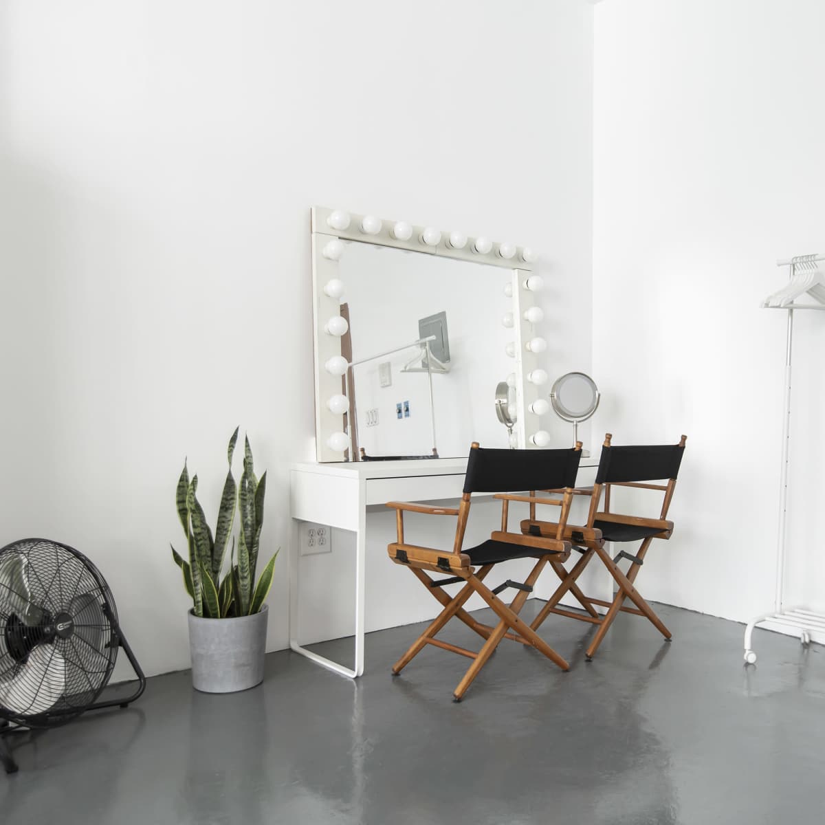 A minimalistic makeup vanity area with two director's chairs, a mirror framed with bulbs, a floor fan, a potted plant, and a clothes rack against a white wall.