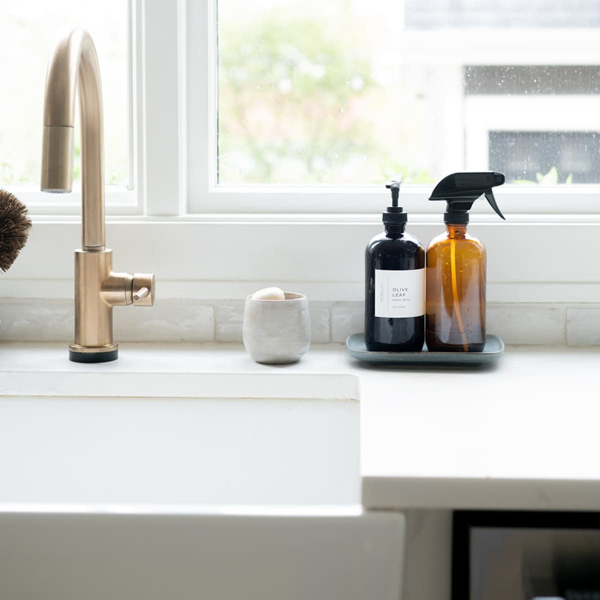A modern kitchen sink area features a bronze faucet, two dark bottles on a tray, and a white ceramic vessel, with natural light streaming through a nearby window.