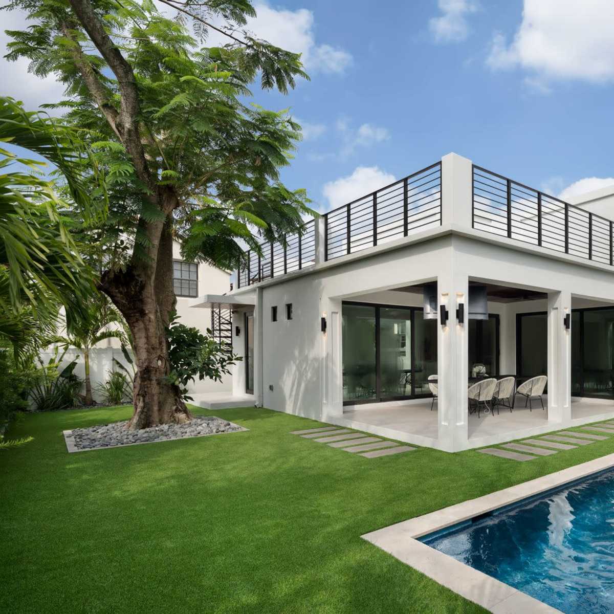 A modern white house with a swimming pool and a landscaped garden featuring green grass and tropical trees.