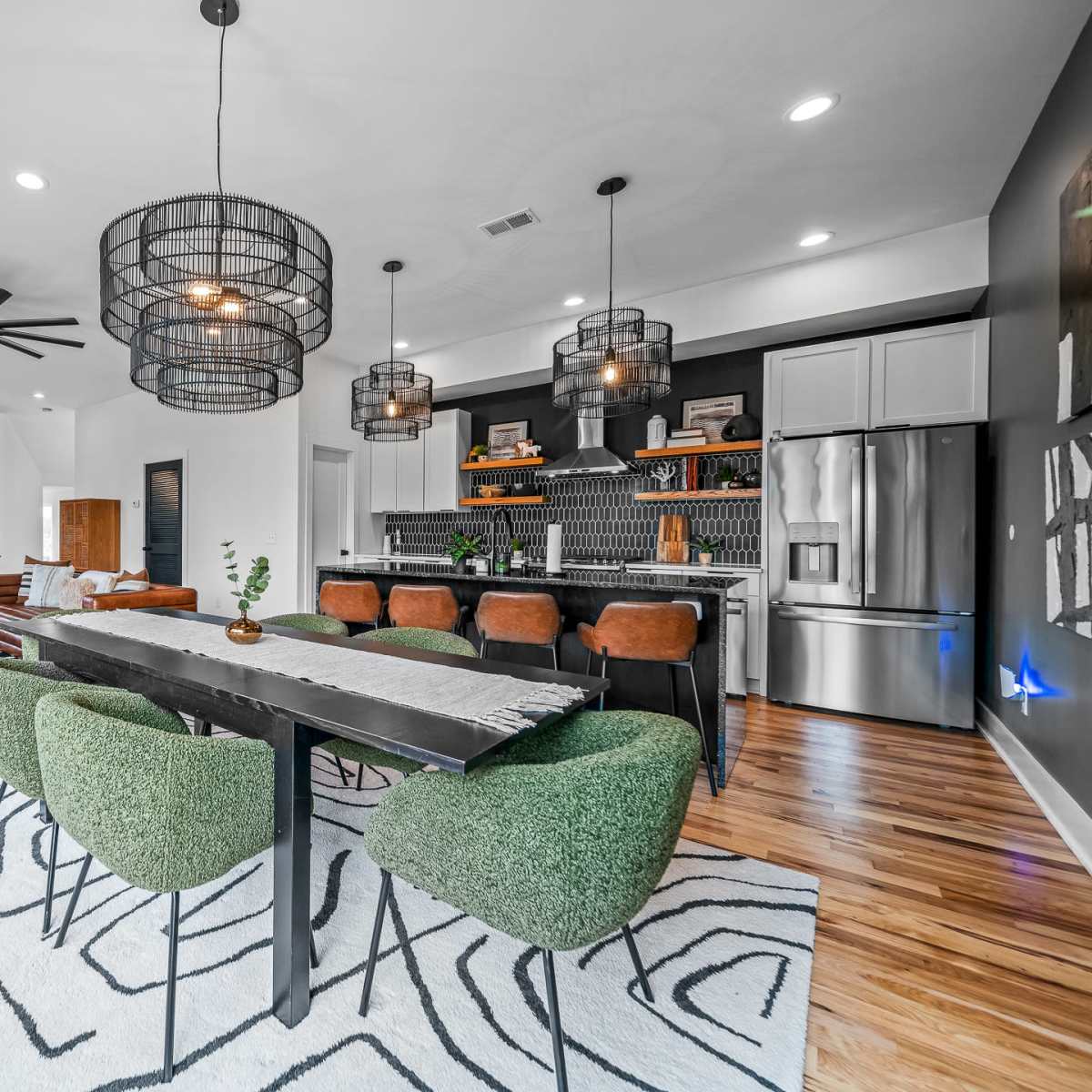A modern open-concept living and dining area featuring a large dining table with green chairs, pendant lights, and a stylish kitchen with stainless steel appliances.