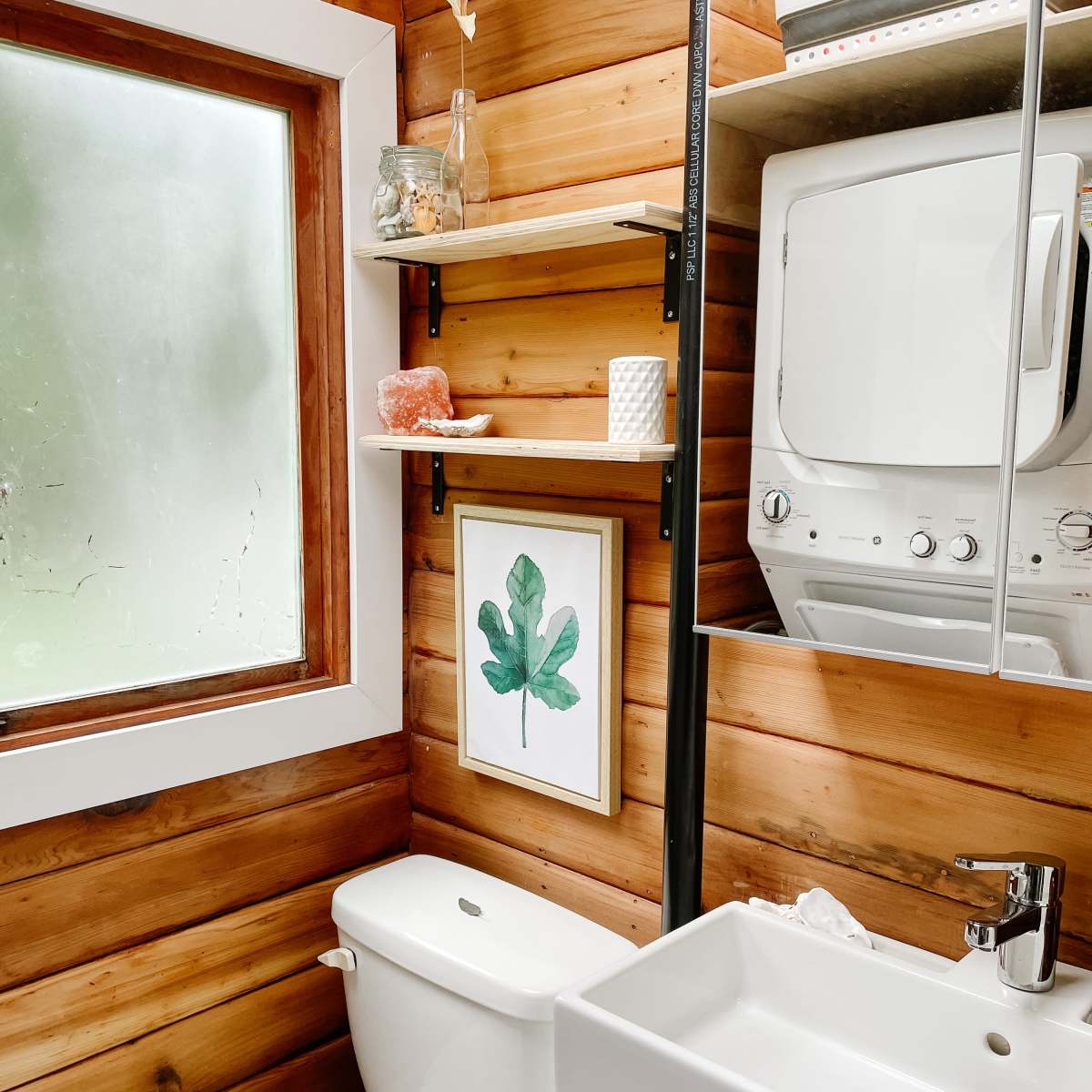 A small bathroom features wooden walls, a wall-mounted sink, a modern toilet, and shelves with decorative items and a framed leaf print.