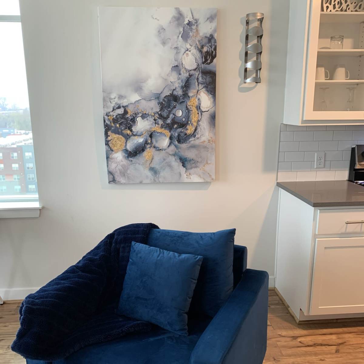 A blue upholstered chair with a navy throw and pillows is positioned next to a wall featuring an abstract painting in shades of gray and gold.