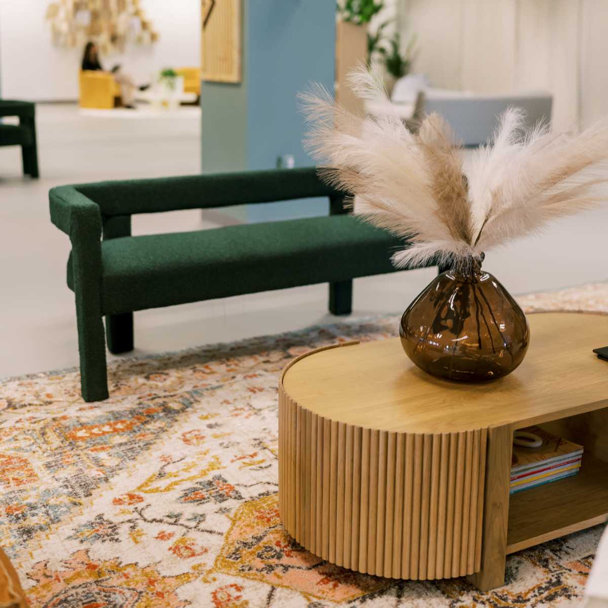 A modern interior space featuring a green sofa and a circular wooden coffee table with a decorative vase and pampas grass, set against a backdrop of neutral-colored walls and furnishings.