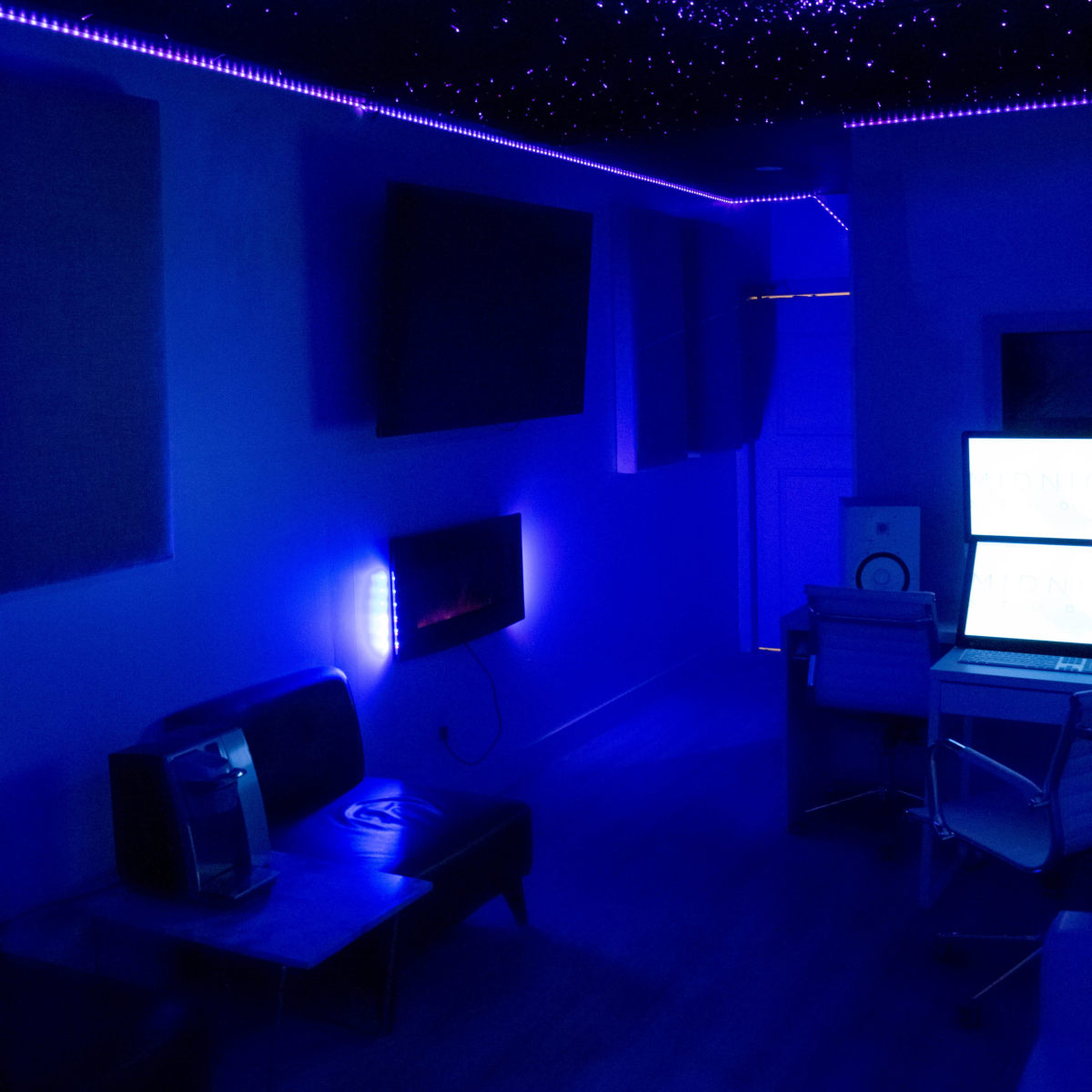 A dimly lit room with blue lighting, featuring a sofa, a television mounted on the wall, and a desk with a computer.