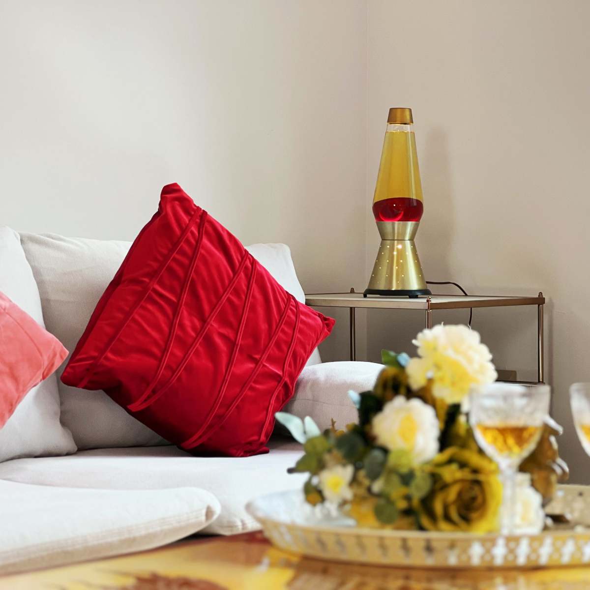 A cozy living room features a white couch with red cushions, a lava lamp on a side table, and a tray with floral decorations and glasses.