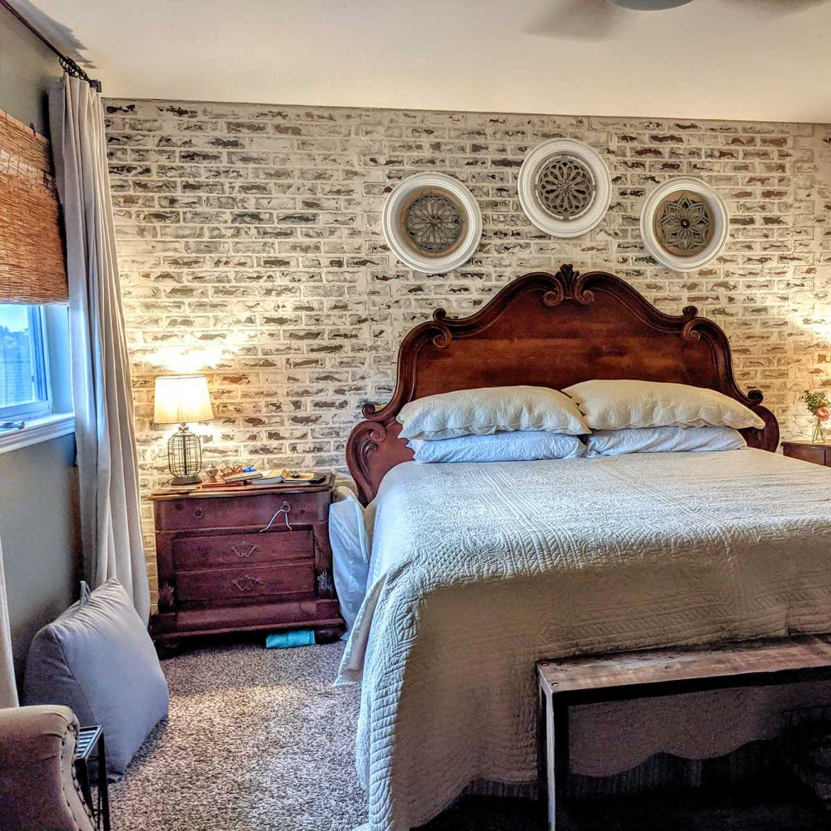 A cozy bedroom features a wooden bed with white bedding, flanked by nightstands and illuminated by lamps, all against a brick-patterned wall.