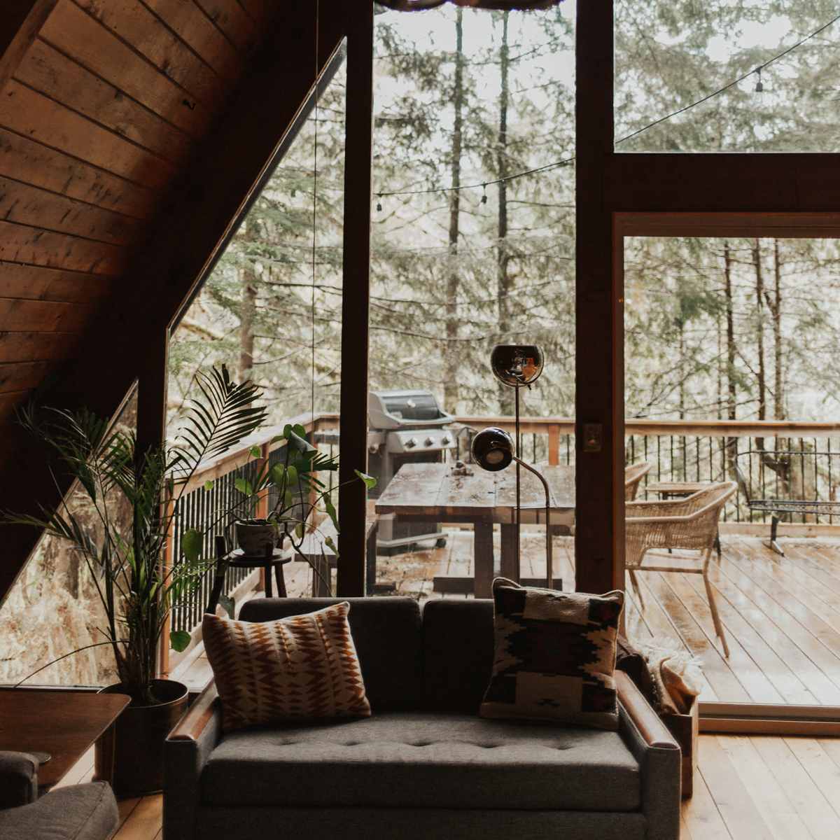 A cozy cabin living area with large windows overlooking a forest, featuring a chandelier made of antlers and a gray sofa.