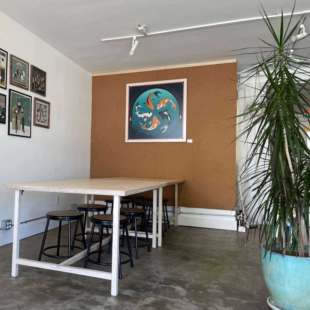 A minimalist room with a large wooden table surrounded by black stools, alongside a wall featuring framed artworks and a corkboard.