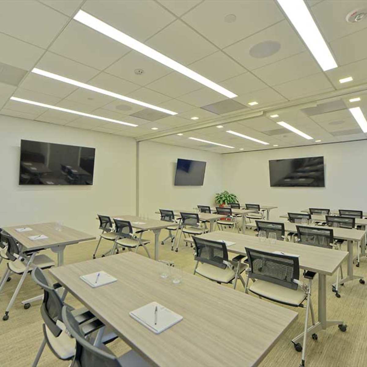 A modern conference room features multiple tables with chairs, two large screens mounted on the walls, and a potted plant in the corner.