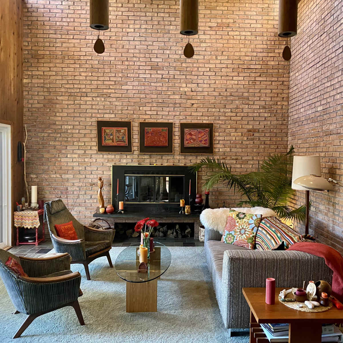 A spacious living room with brick walls, a black fireplace, a gray sofa, and two armchairs, complemented by a round glass coffee table and a large window letting in natural light.