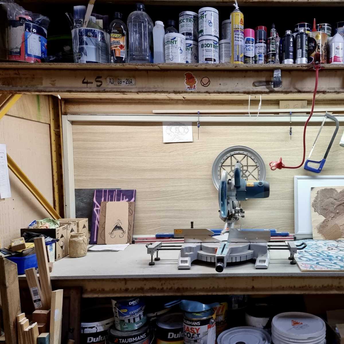 A workshop shelf displays various tools, paint cans, and materials, with a miter saw set up on the workbench.