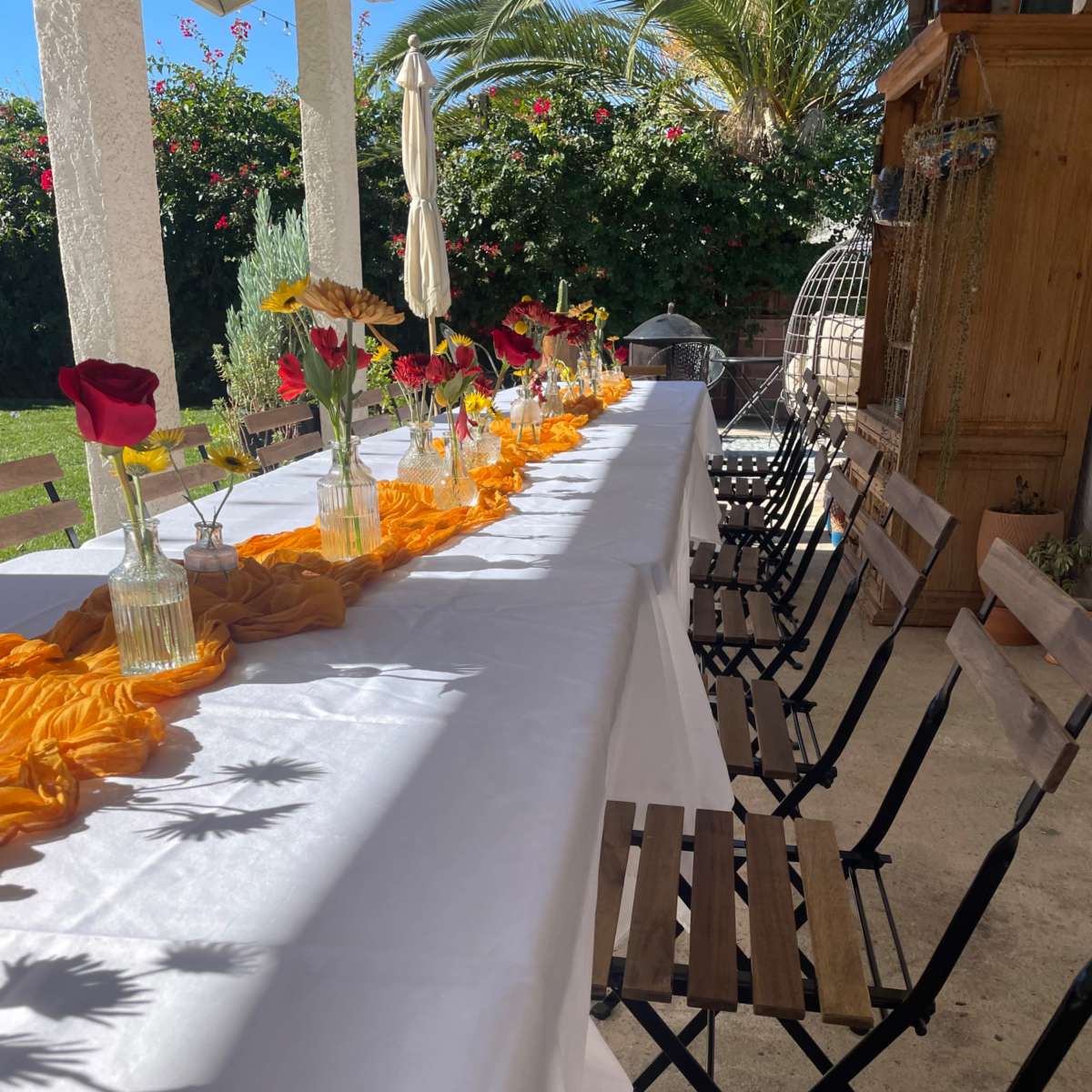 A long table with a white tablecloth is set outdoors, adorned with vases of flowers and a decorative orange cloth, surrounded by wooden chairs.