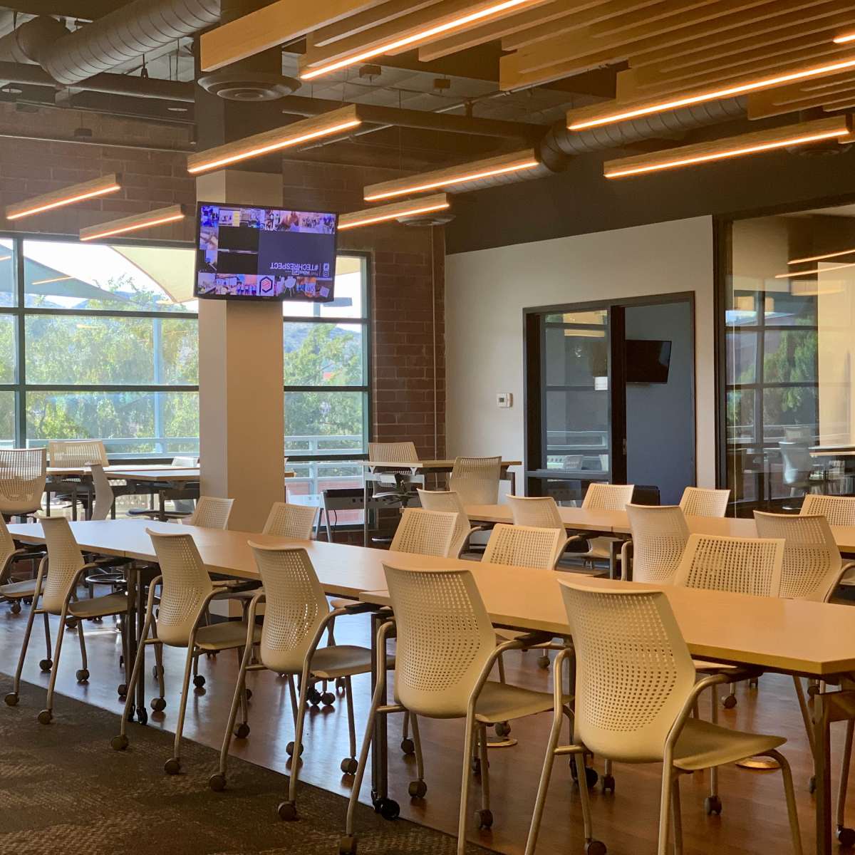 A modern conference room with multiple tables and chairs arranged for meetings, featuring large windows and a television screen mounted on the wall.