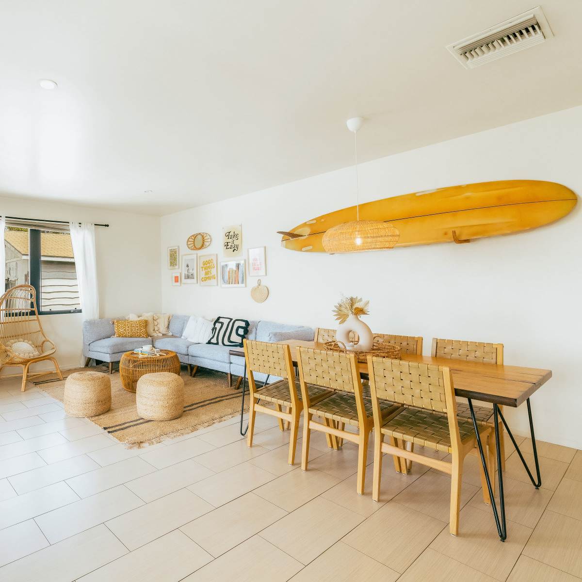 A bright, modern living space featuring a light blue sofa, a wooden dining table with chairs, and surfboard decor on the wall.