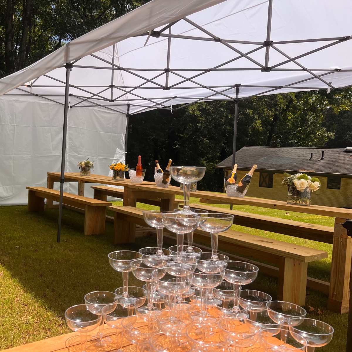 A white canopy is set up outdoors over wooden benches, with a display of champagne glasses and bottles arranged on a table in the background.