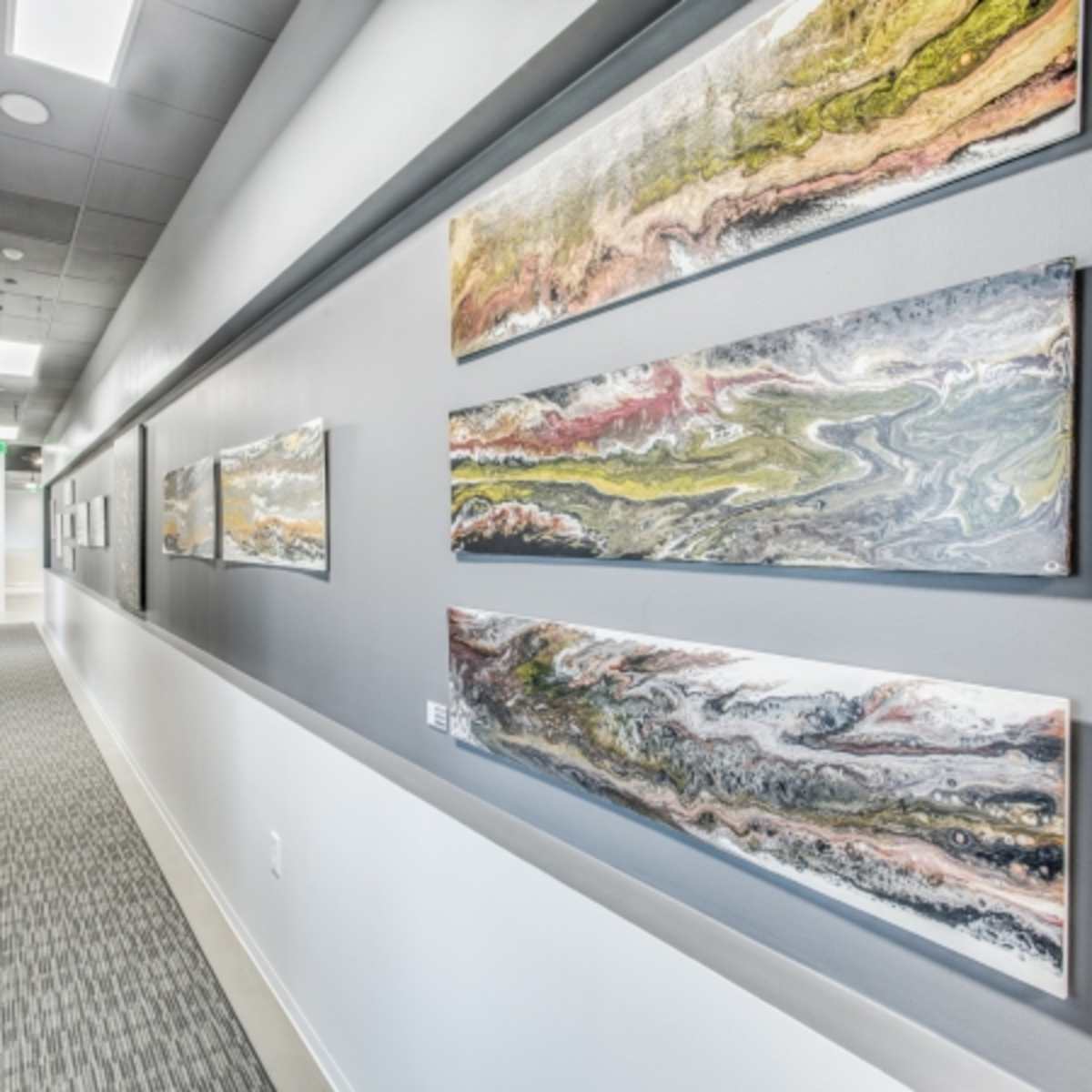 A narrow hallway features gray walls adorned with three large abstract paintings and a patterned carpet.