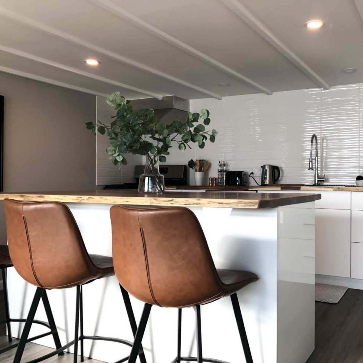 A modern kitchen features a wooden countertop island with brown bar stools, white cabinetry, and stainless steel appliances.