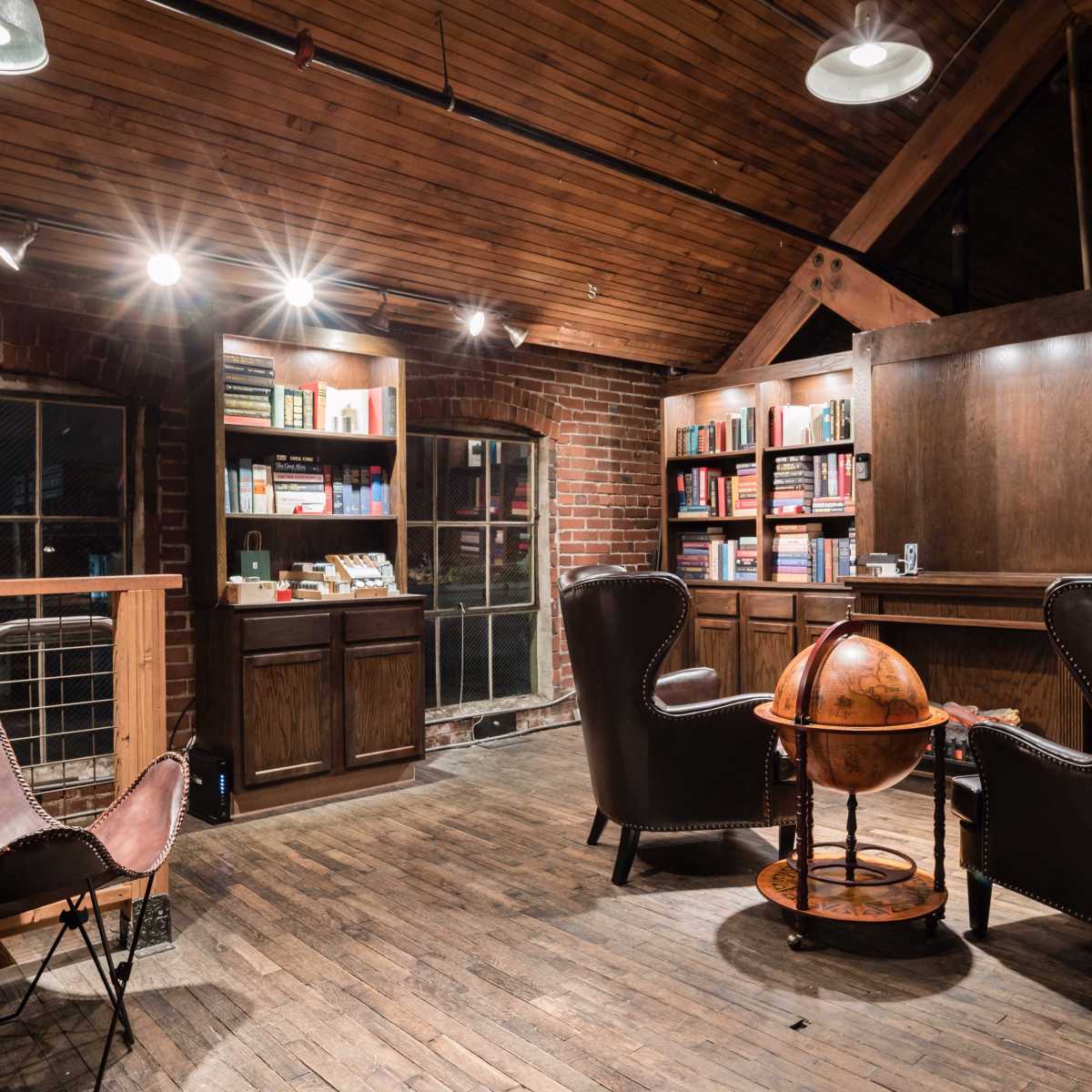 A cozy library with wooden shelves filled with books, two leather chairs, and a globe on a wooden stand, all set in a space featuring brick walls and a wooden ceiling.