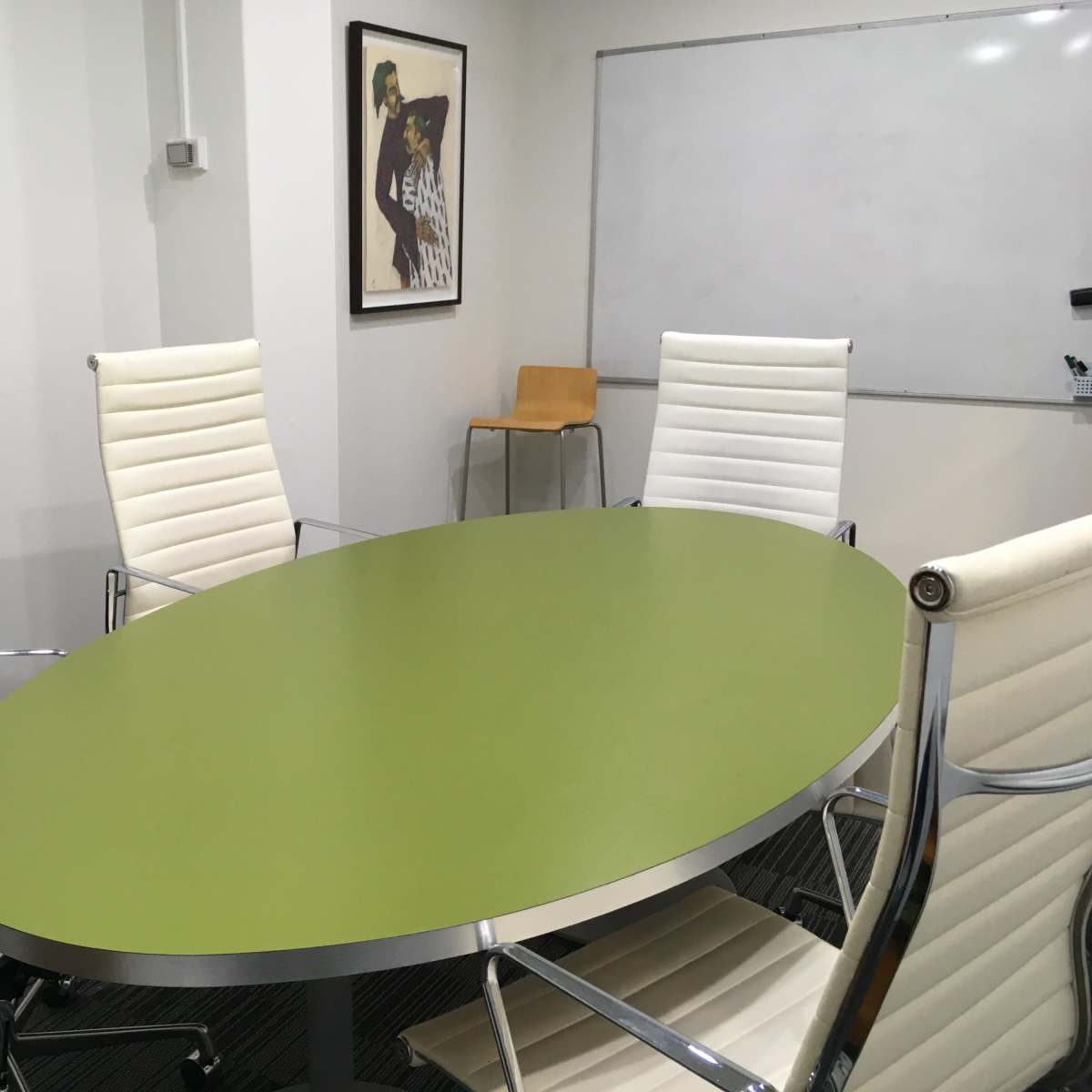 A modern conference room features a green oval table surrounded by white leather chairs, with a framed artwork on the wall and a whiteboard in the background.
