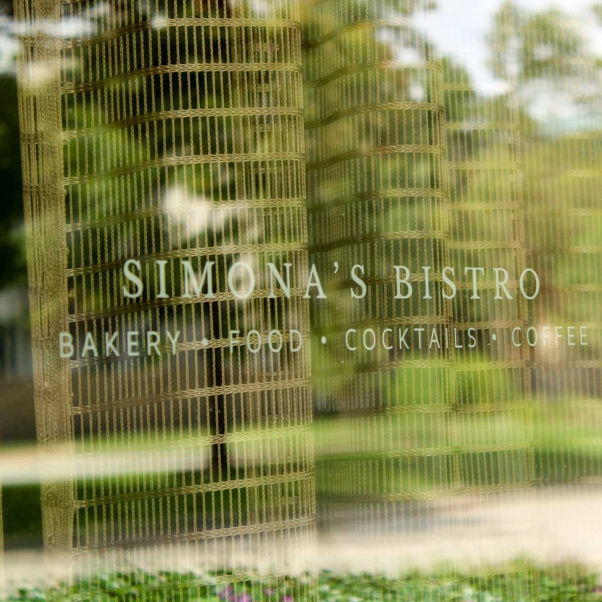 A reflective glass surface displaying the sign for "Simona's Bistro," which offers bakery items, food, cocktails, and coffee.
