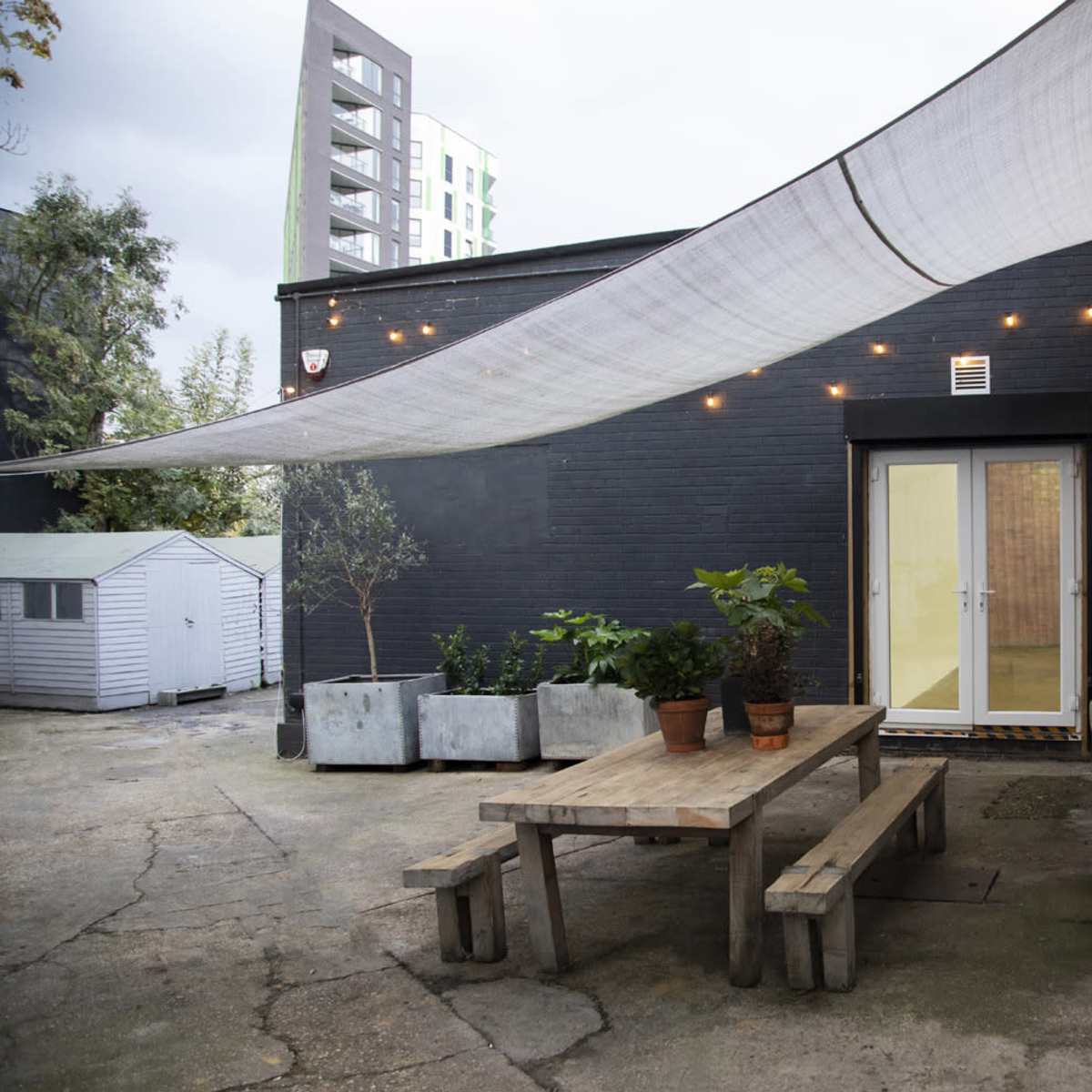 A wooden table with benches is positioned under a large fabric canopy and string lights, next to a black wall and several potted plants.