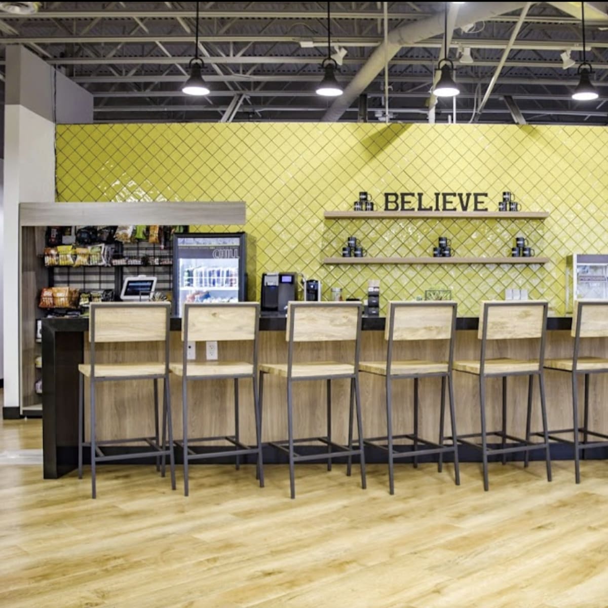 A modern café bar area with wooden high chairs facing a counter against a yellow patterned wall featuring the word "BELIEVE."
