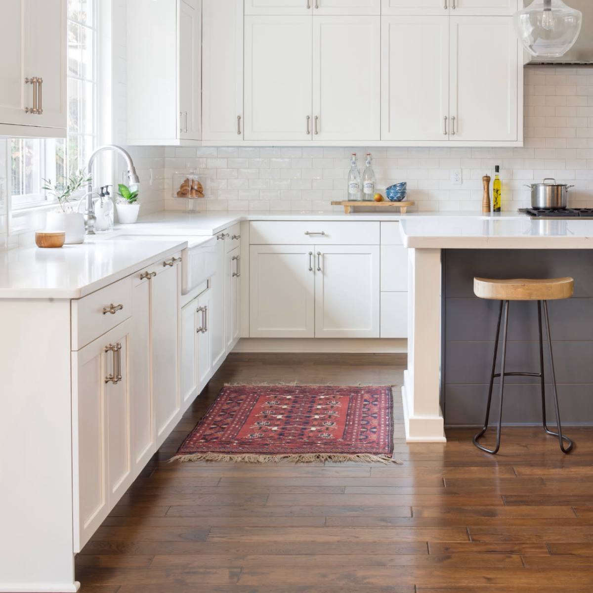 A bright kitchen features white cabinets, a marble countertop, and wooden flooring, with a decorative rug and modern bar stool.