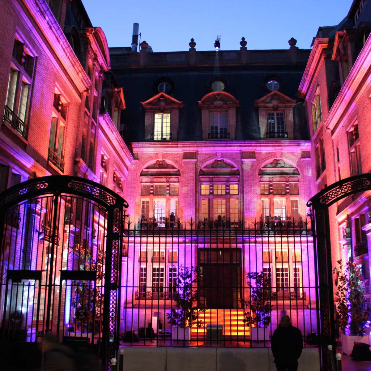 A historic building with illuminated pink and purple lighting, surrounded by a gated entrance and trees at dusk.