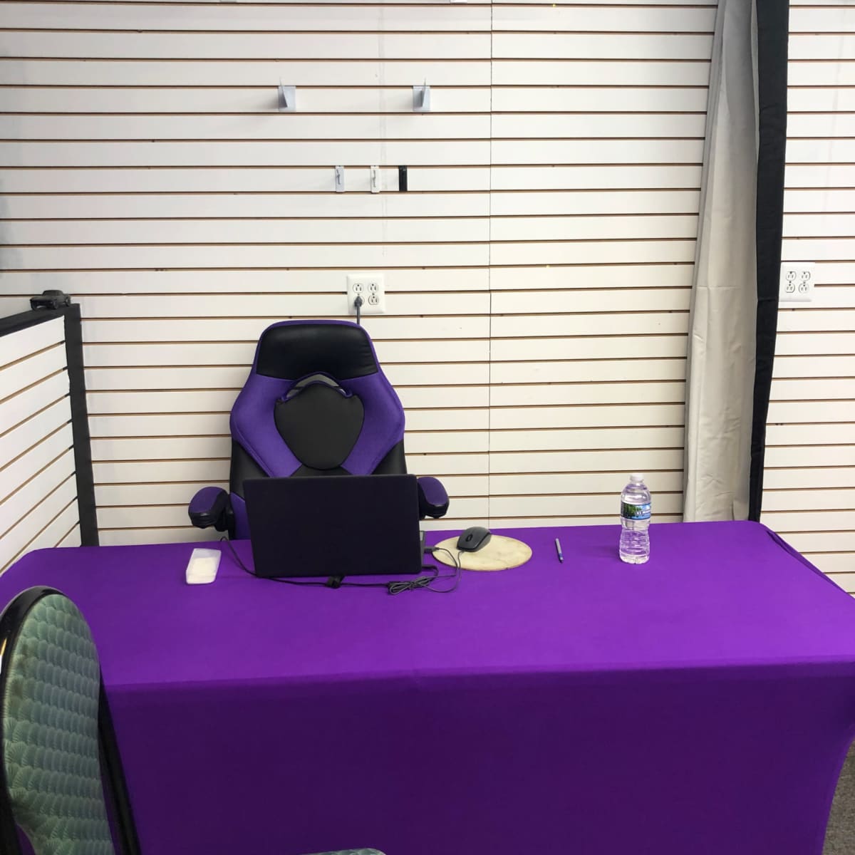 A purple desk with a computer and a water bottle is set up in front of a purple gaming chair against a slatted wall.