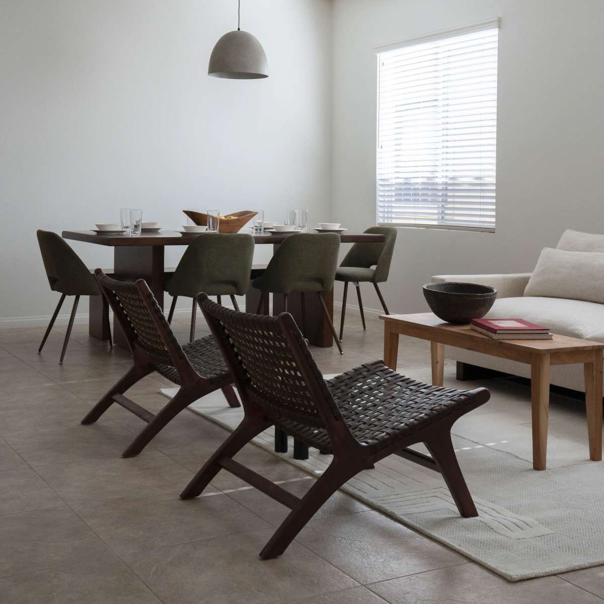 A modern, minimalistic living space features a dining table with chairs, two woven lounge chairs, and a sofa, all arranged on tiled flooring.