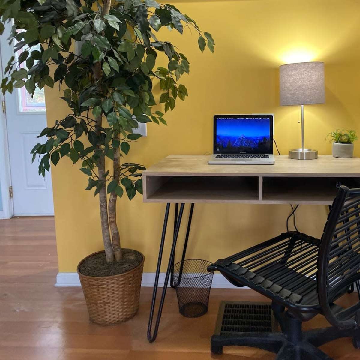 A small workspace features a wooden desk with a laptop, a lamp, a potted plant, and a black chair, set against a yellow wall next to a decorative tree.